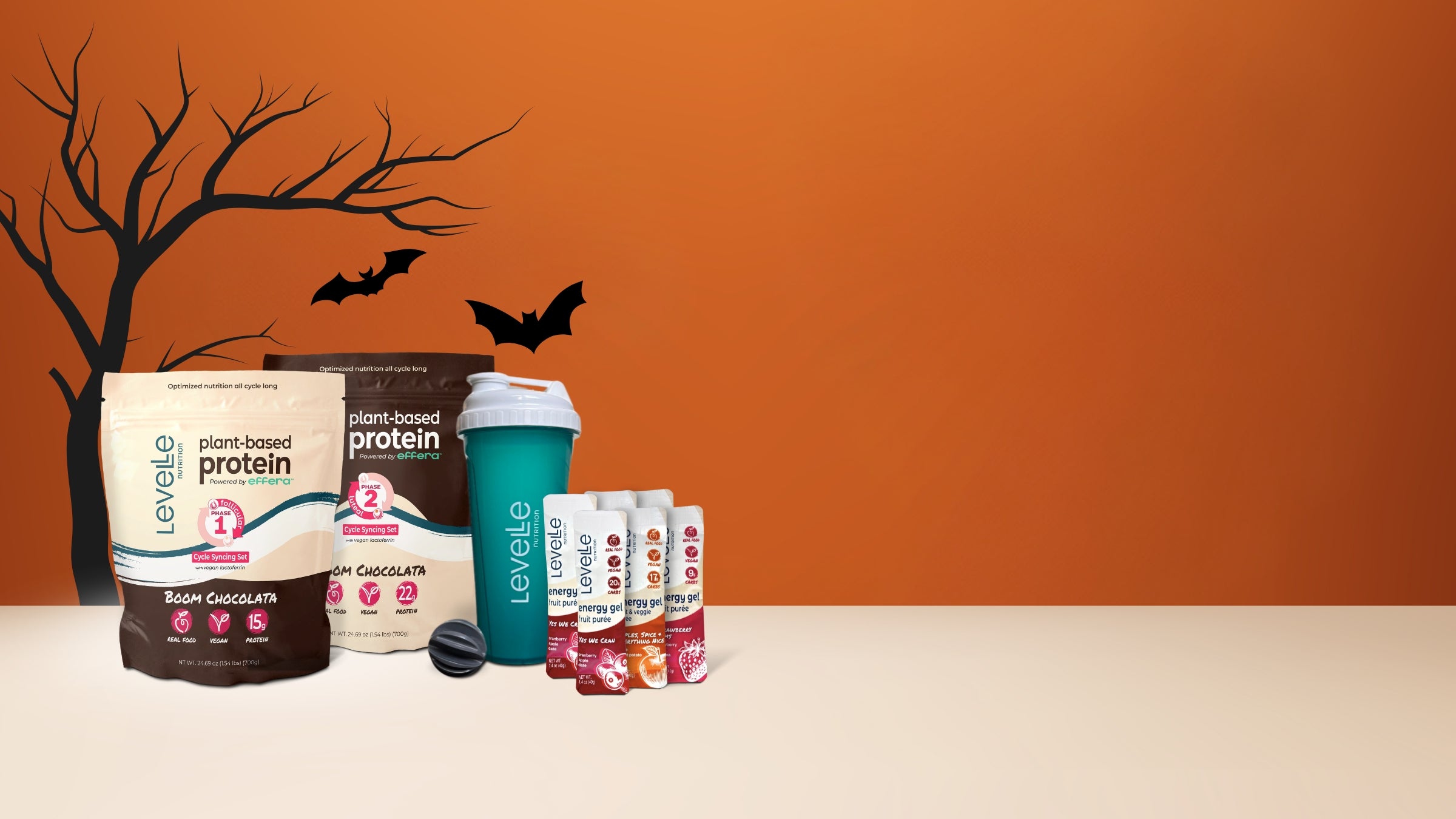 Protein powder packages, energy gels and a shaker cup on a table with an orange wall and black bats in the background.