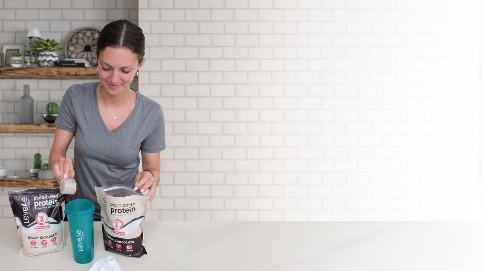 Woman in a kitchen preparing a protein shake with a bag of protein powder and a shaker bottle.